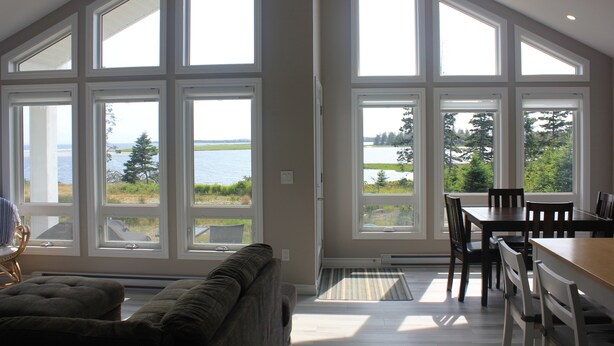 A spacious room with large windows offering a view of a lake and trees. There is a dining area with a table and chairs. - Cottage at Cherry Hill Beach (Cherry Hill)