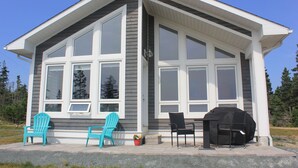 A modern house with large windows, a porch with two chairs, and a barbecue grill. - Cottage at Cherry Hill Beach (Cherry Hill)