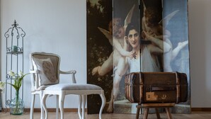 Ein Zimmer mit einem weißen Polsterstuhl, einem alten Koffer und einem großen Gemälde von Cherubs an der Wand.