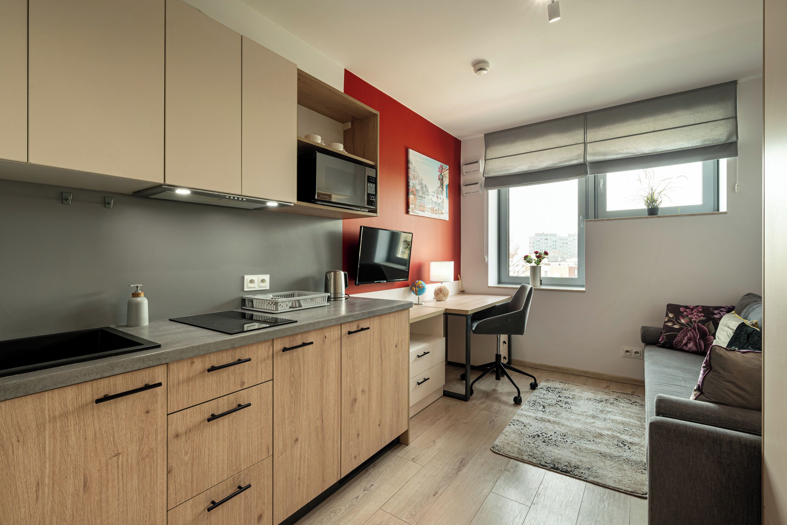 A modern kitchen with wooden cabinets, a microwave, and a built-in oven. There's a workspace with a desk and chair, and a window with a view of buildings.
