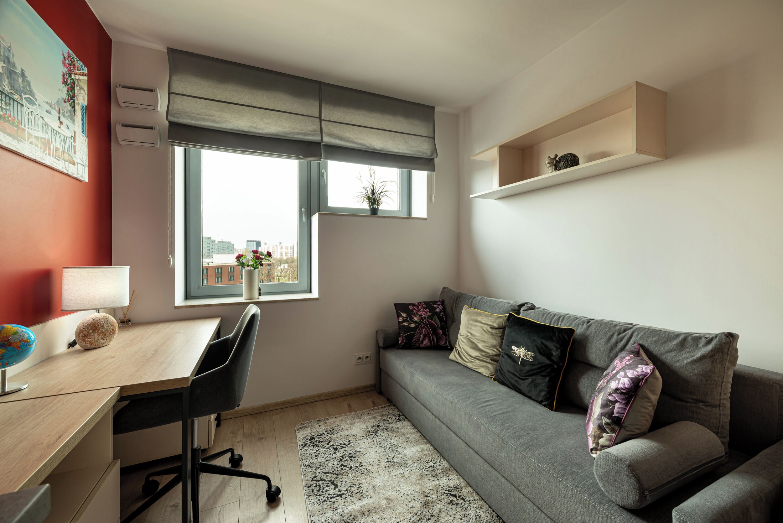 A modern room with a desk, a grey sofa, a window with a view, and a shelf with a plant.