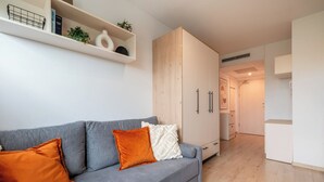 A modern living room with a grey sofa, orange cushions, a wooden wardrobe, and a white shelf with books and plants. - RentPlanet - Apartamenty Dyrekcyjna (Wroclaw)