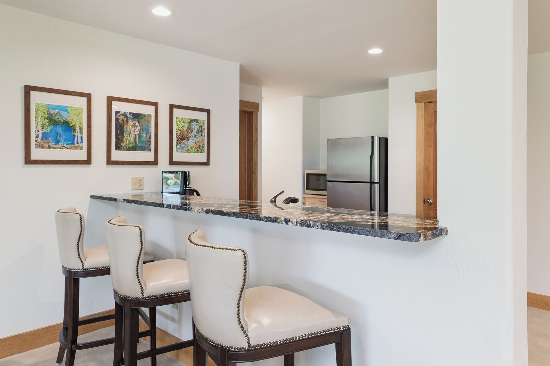 A kitchen with a marble countertop, three bar stools, a refrigerator, and a television.
