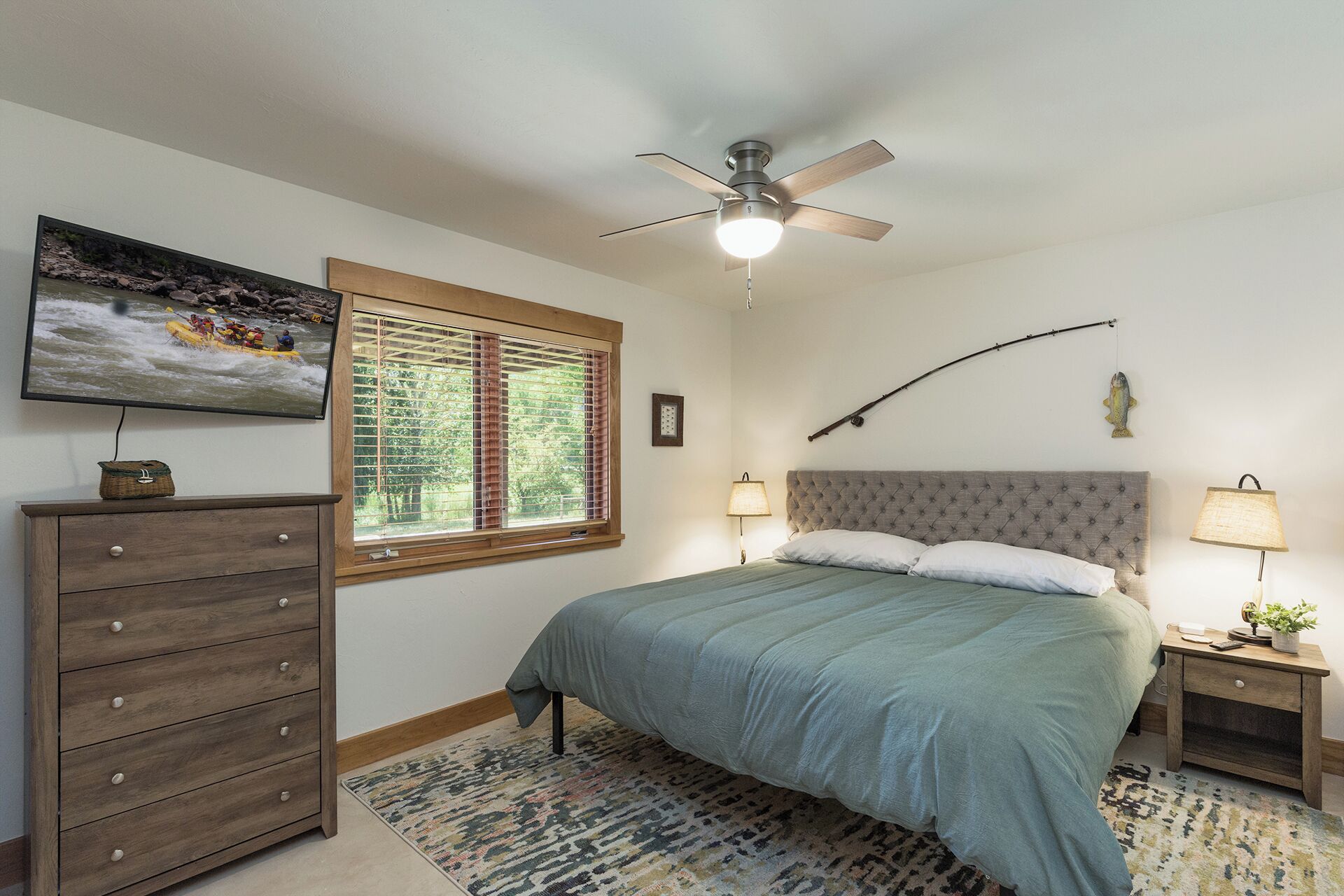 A bedroom with a bed, a dresser, a ceiling fan, a window with blinds, a painting, and a lamp on a bedside table.