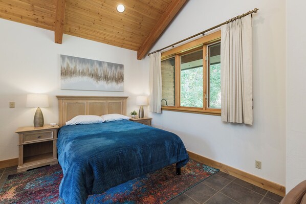 Een slaapkamer met een houten plafond, een groot bed met een blauwe plaid, een nachtkastje met een lamp, een schilderij aan de muur en een raam met gordijnen.