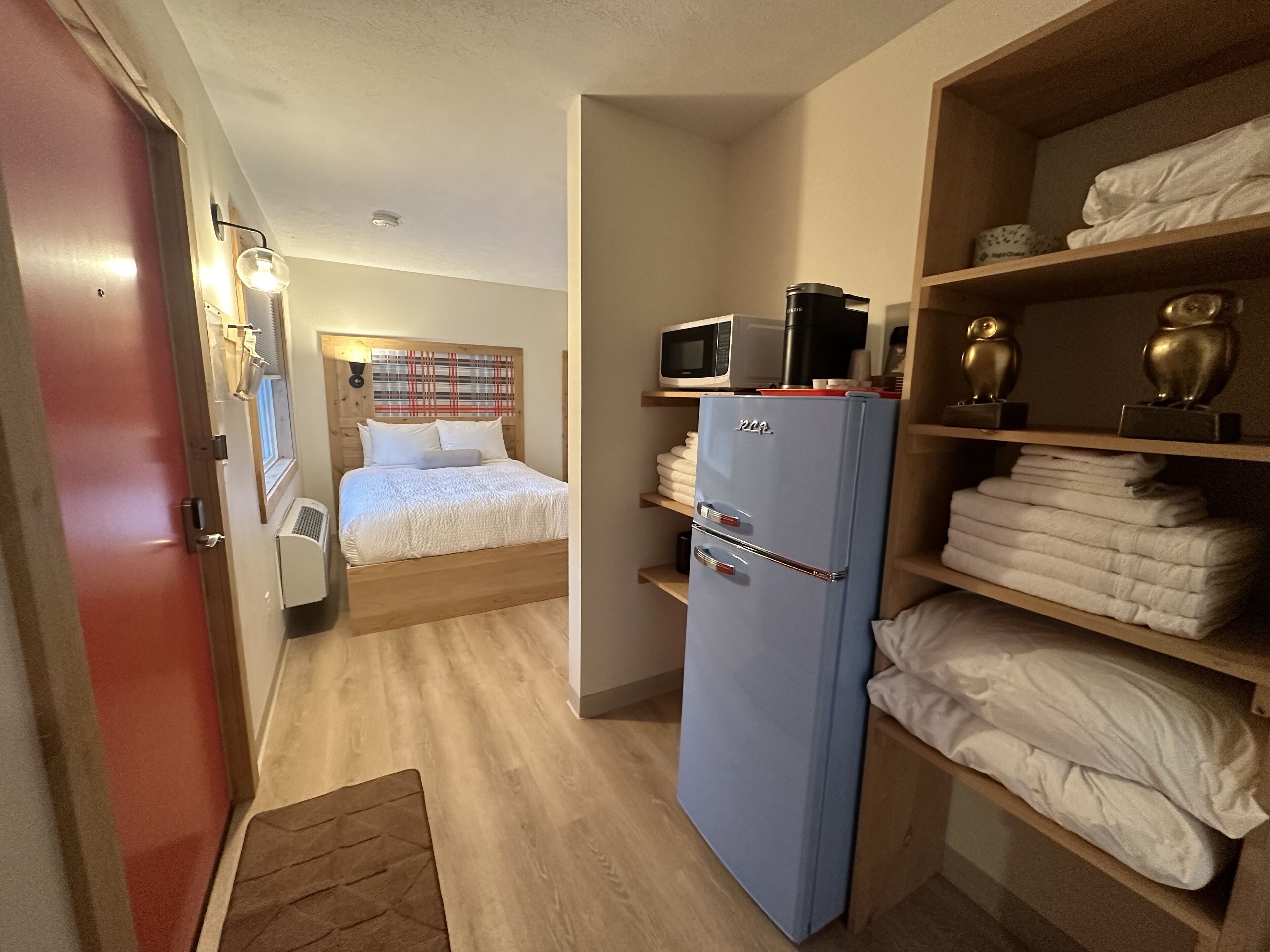 A compact hotel room with a bed, a kitchenette with a microwave, a refrigerator, and shelves with towels.