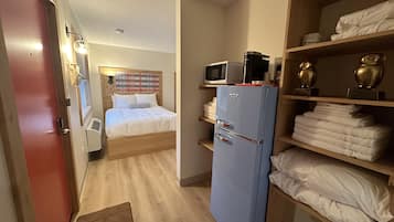 A compact hotel room with a bed, a kitchenette with a microwave, a refrigerator, and shelves with towels.
