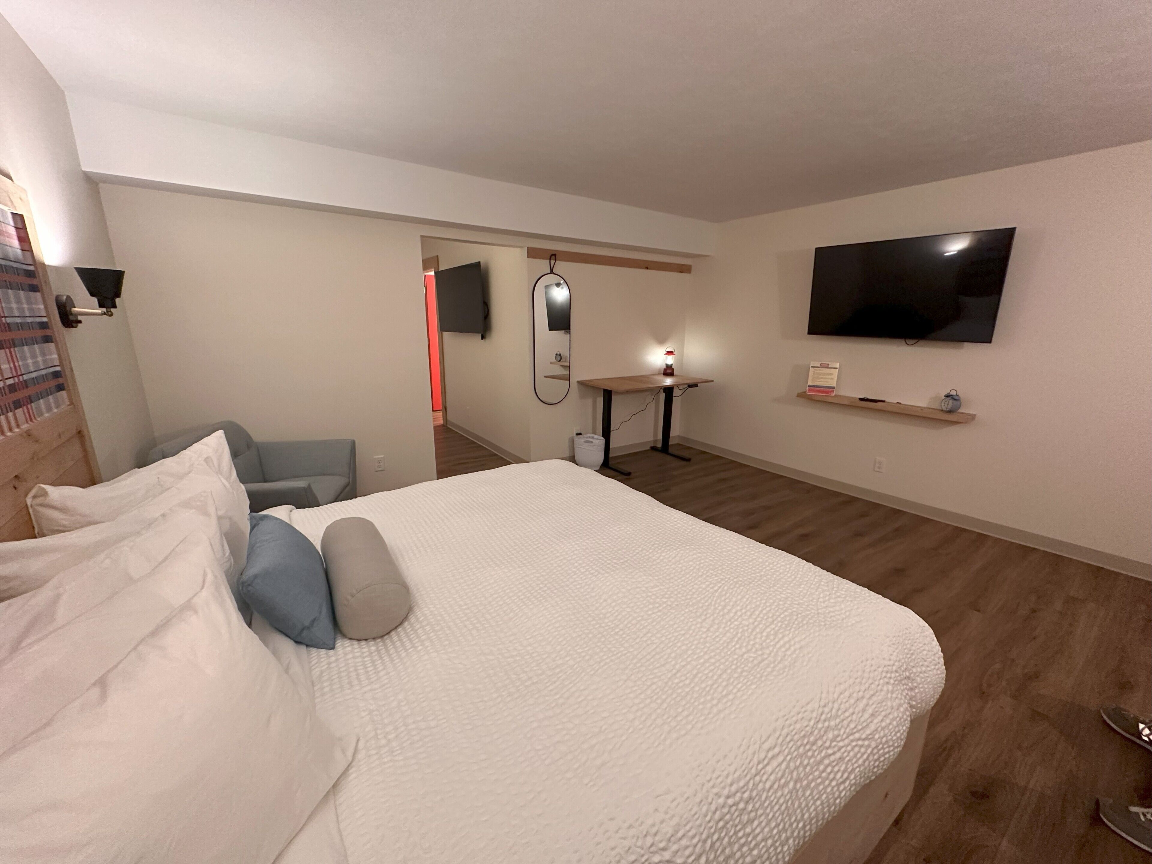A hotel room with a bed, a desk, a chair, a TV, and a small shelf.