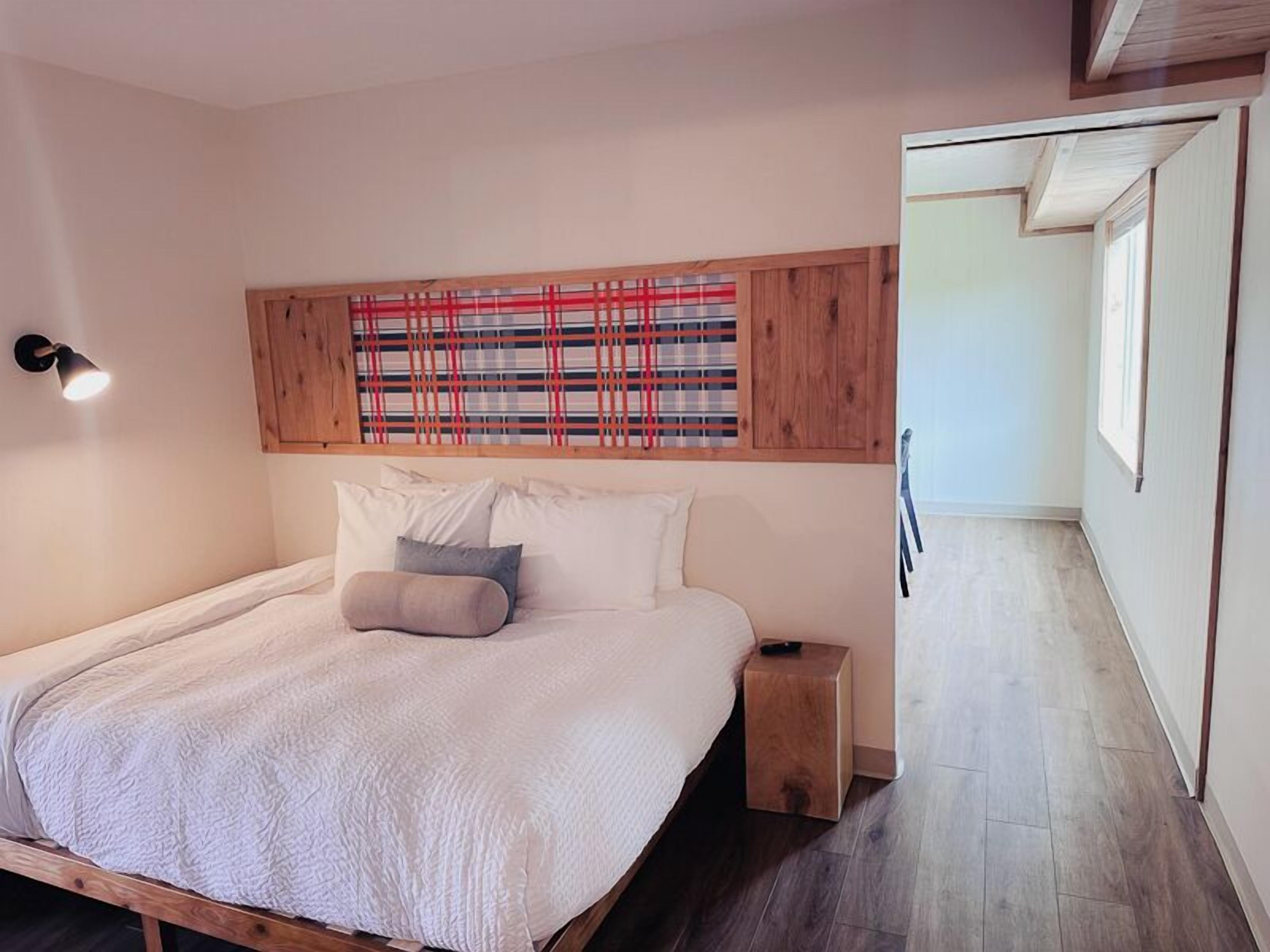 A bedroom with a wooden headboard, a bed with white bedding, a bedside table, and a view of a hallway.