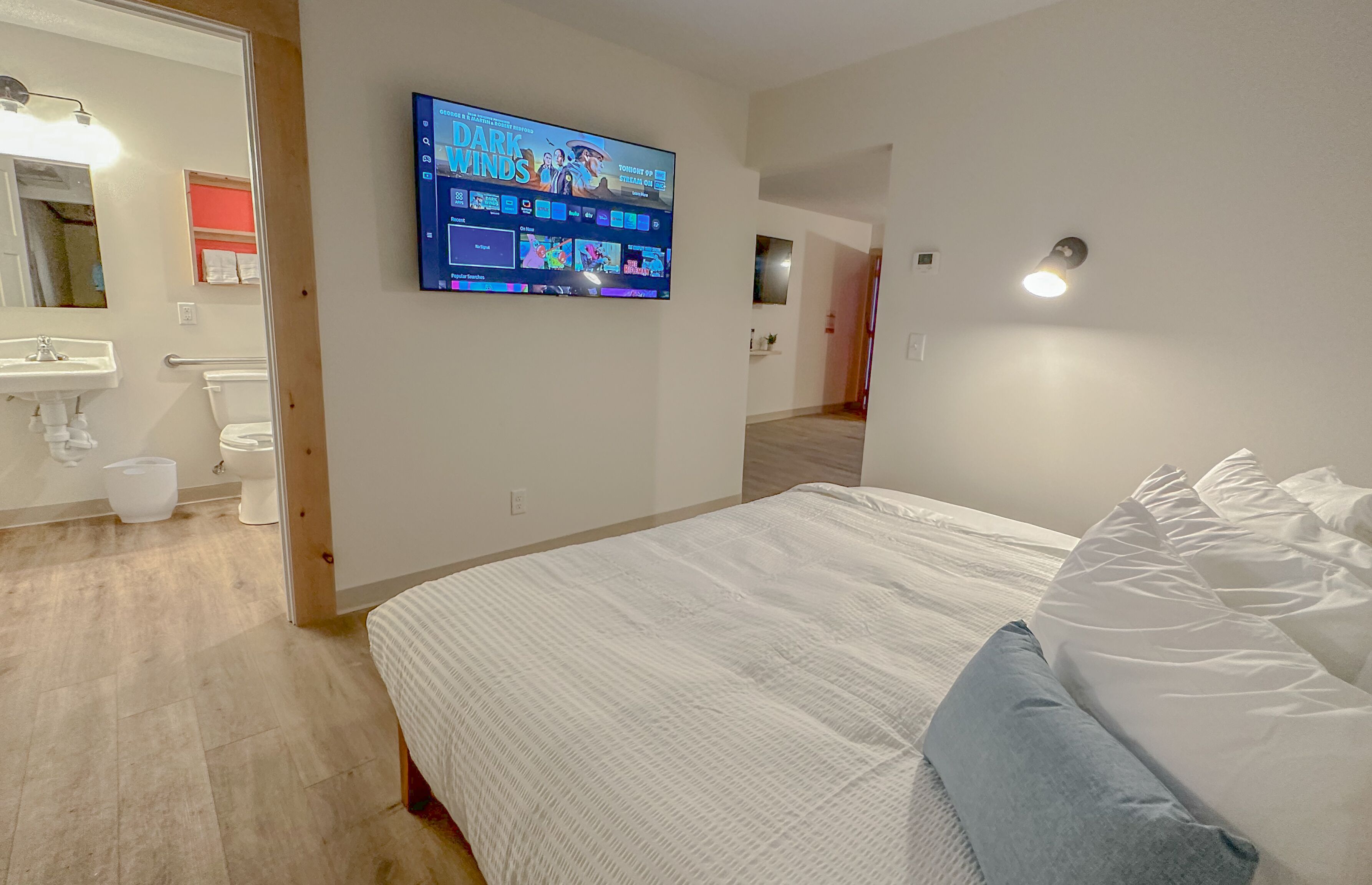 A bedroom with a bed, a TV mounted on the wall, a bathroom with a toilet and sink, and a hallway leading to another room.