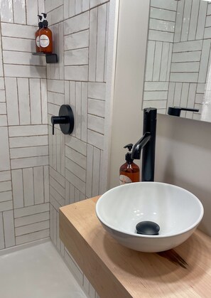 A bathroom with a white tiled shower wall, a wooden countertop, a white sink with a black faucet, and a bottle of shampoo and soap.