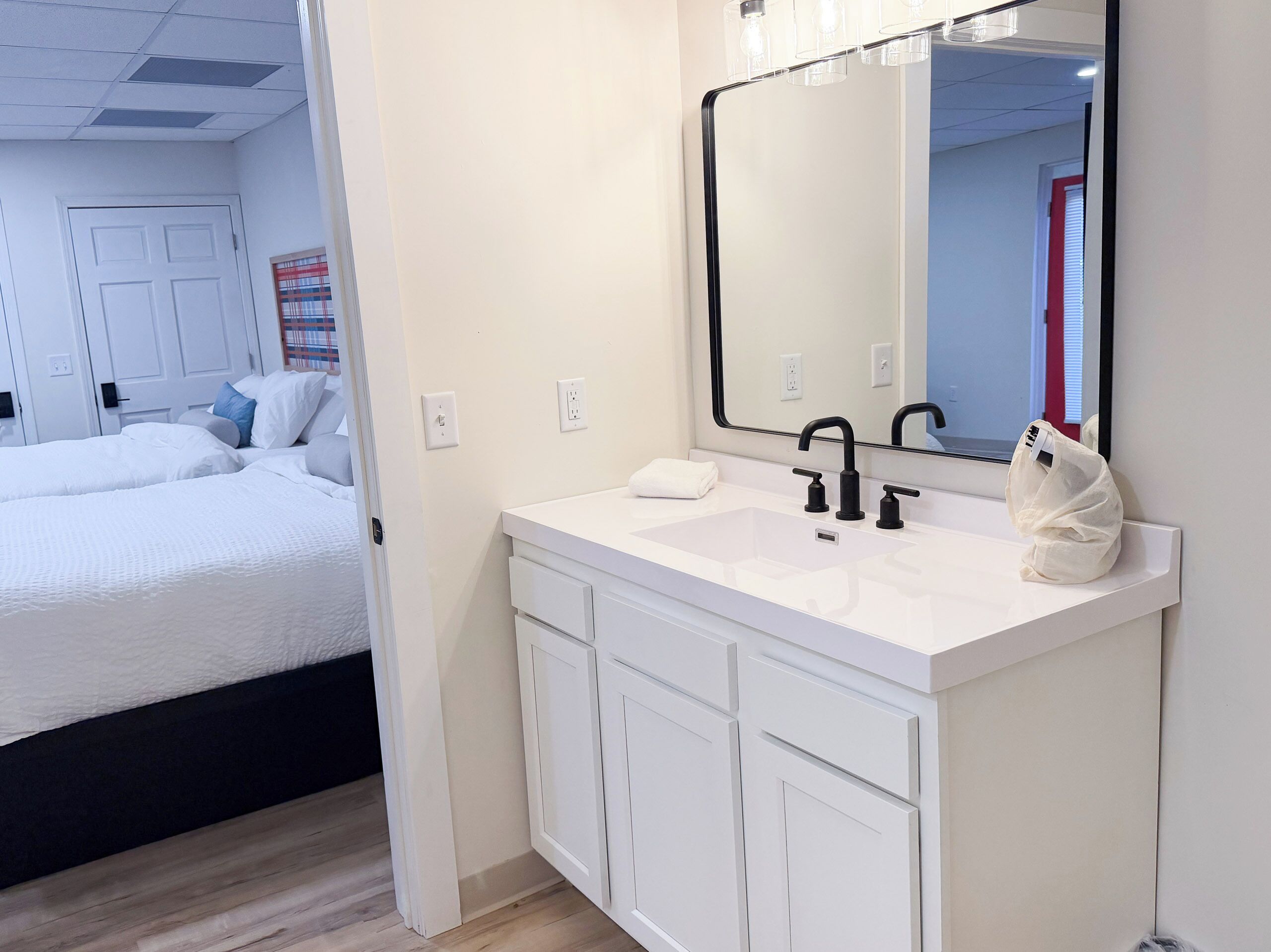 A modern bathroom with a white sink, black faucet, and a large mirror. There is a white towel on the sink and a white bag on the countertop.