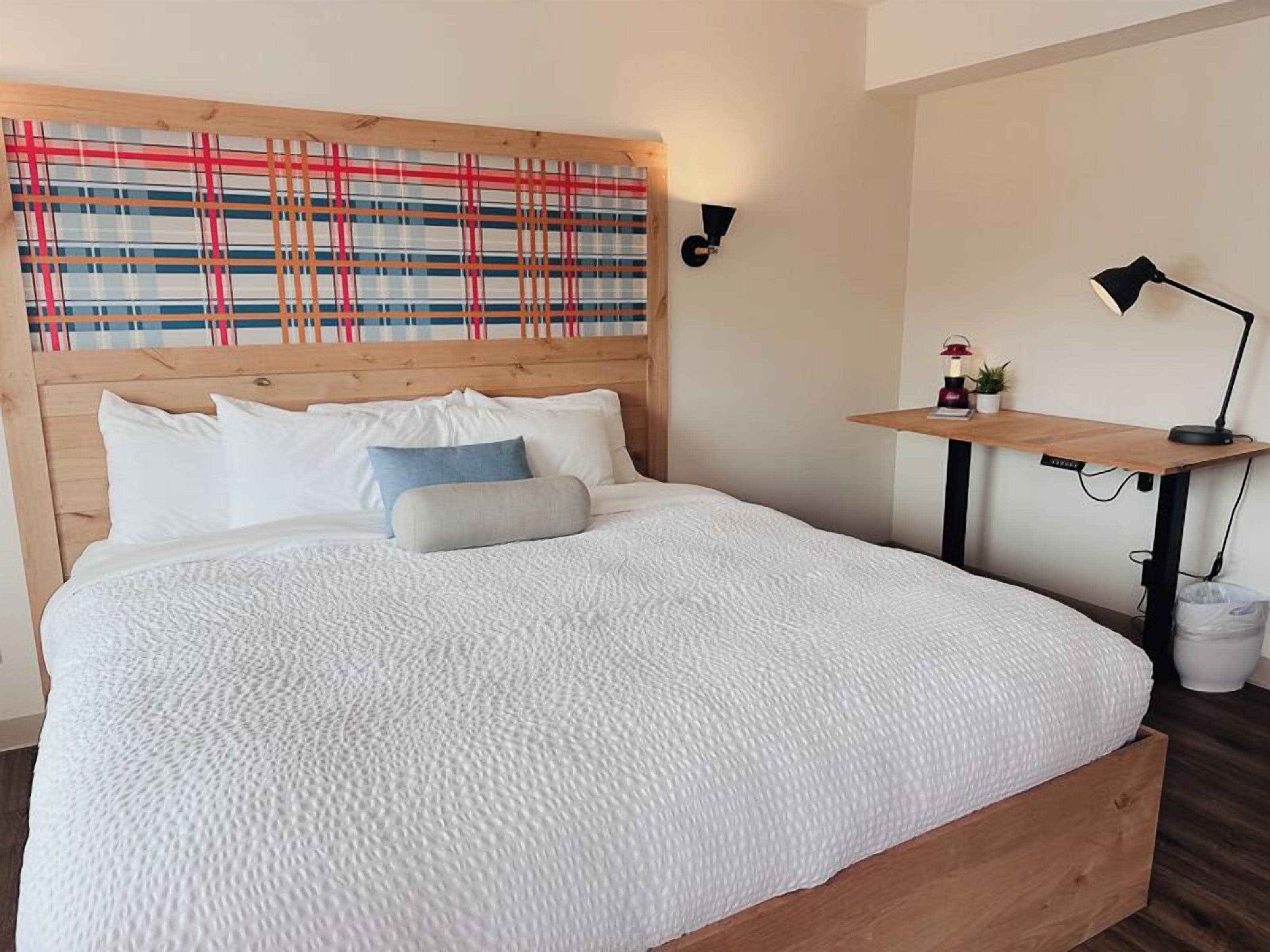 A neatly made bed with white bedding, a wooden headboard, and a plaid-patterned wall hanging above.