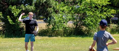 En person i en sort trøje og turkis shorts spiller baseball på en græsplæne med træer i baggrunden.