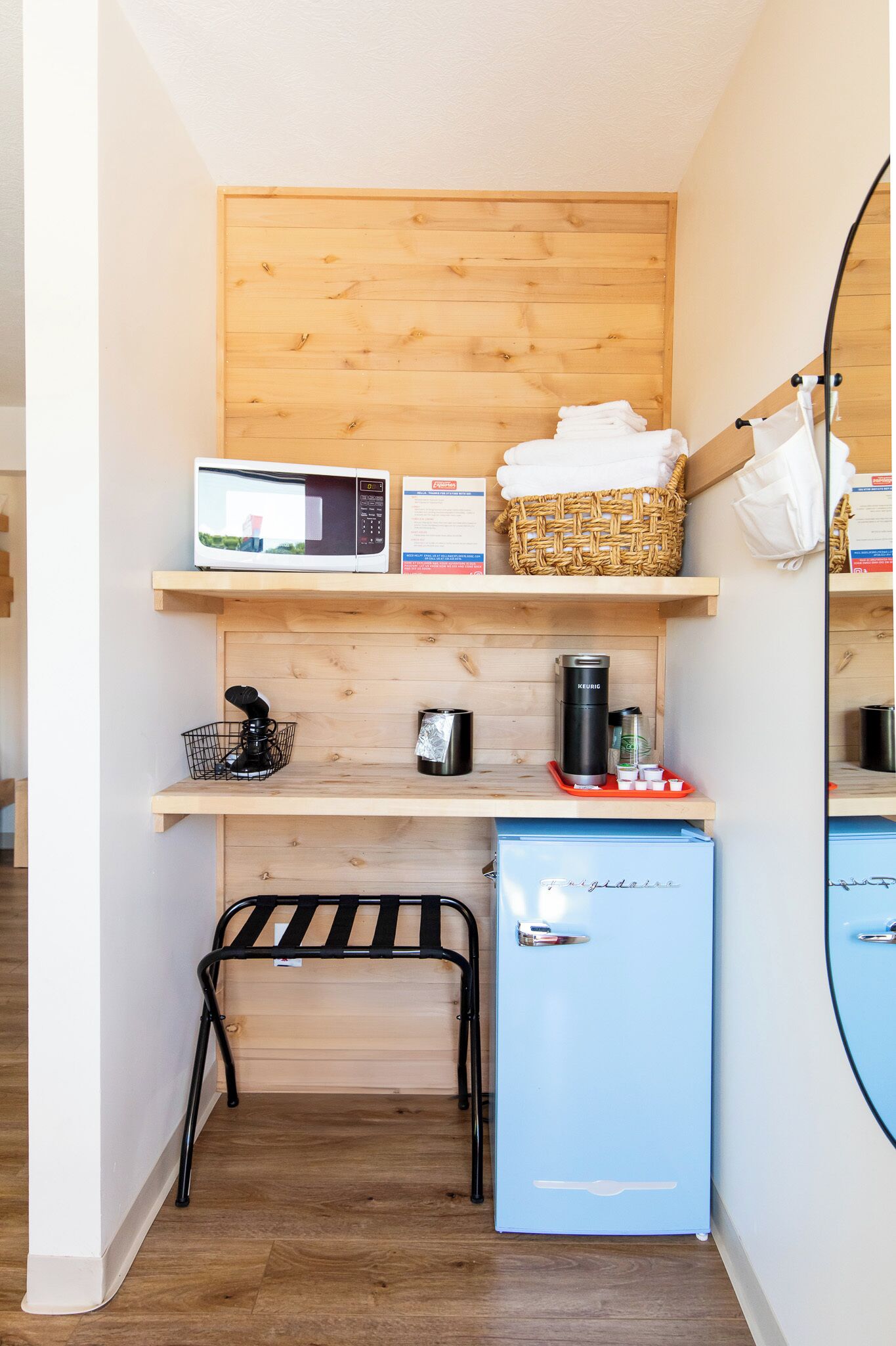 A compact kitchenette with a microwave, a small fridge, a coffee maker, and a basket on a wooden shelf.