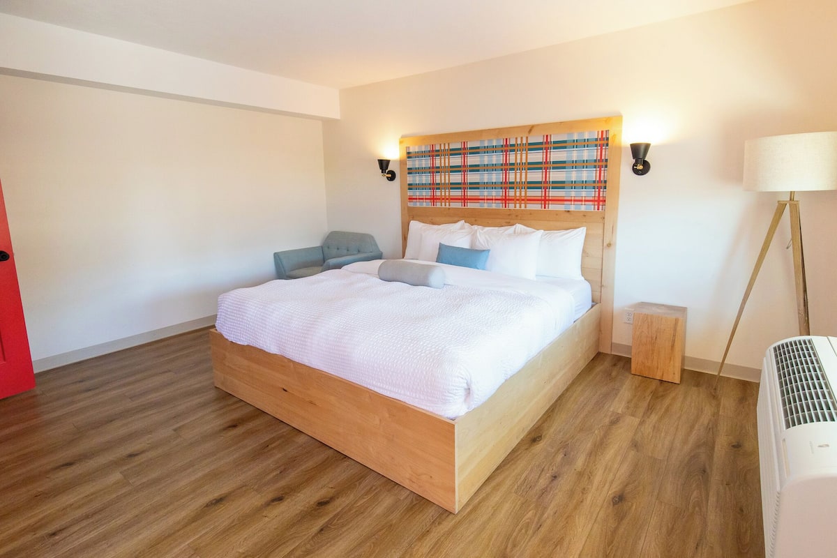 Quarto moderno com cama de madeira, cabeceira com padrão colorido, mesa de cabeceira, abajur e porta vermelha.