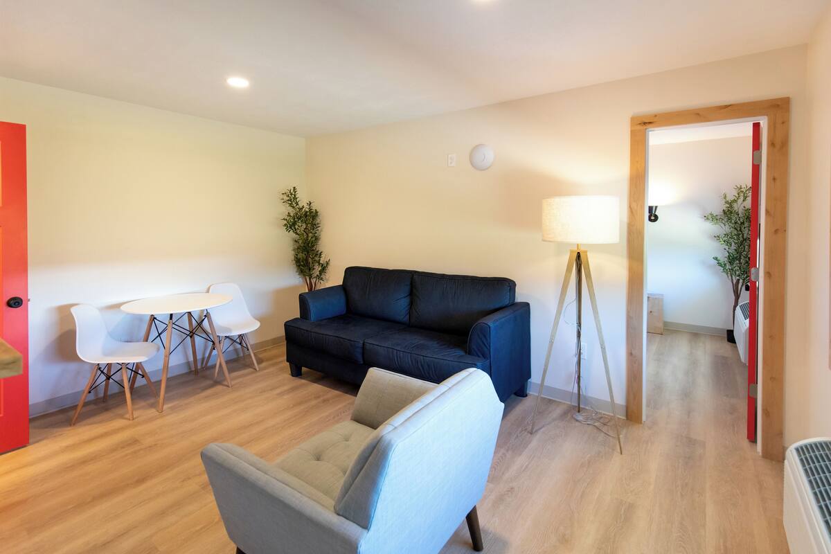 Uma sala de estar com um sofá azul escuro, uma poltrona cinza claro, uma mesa branca com quatro cadeiras brancas, um abajur e um vaso de planta.