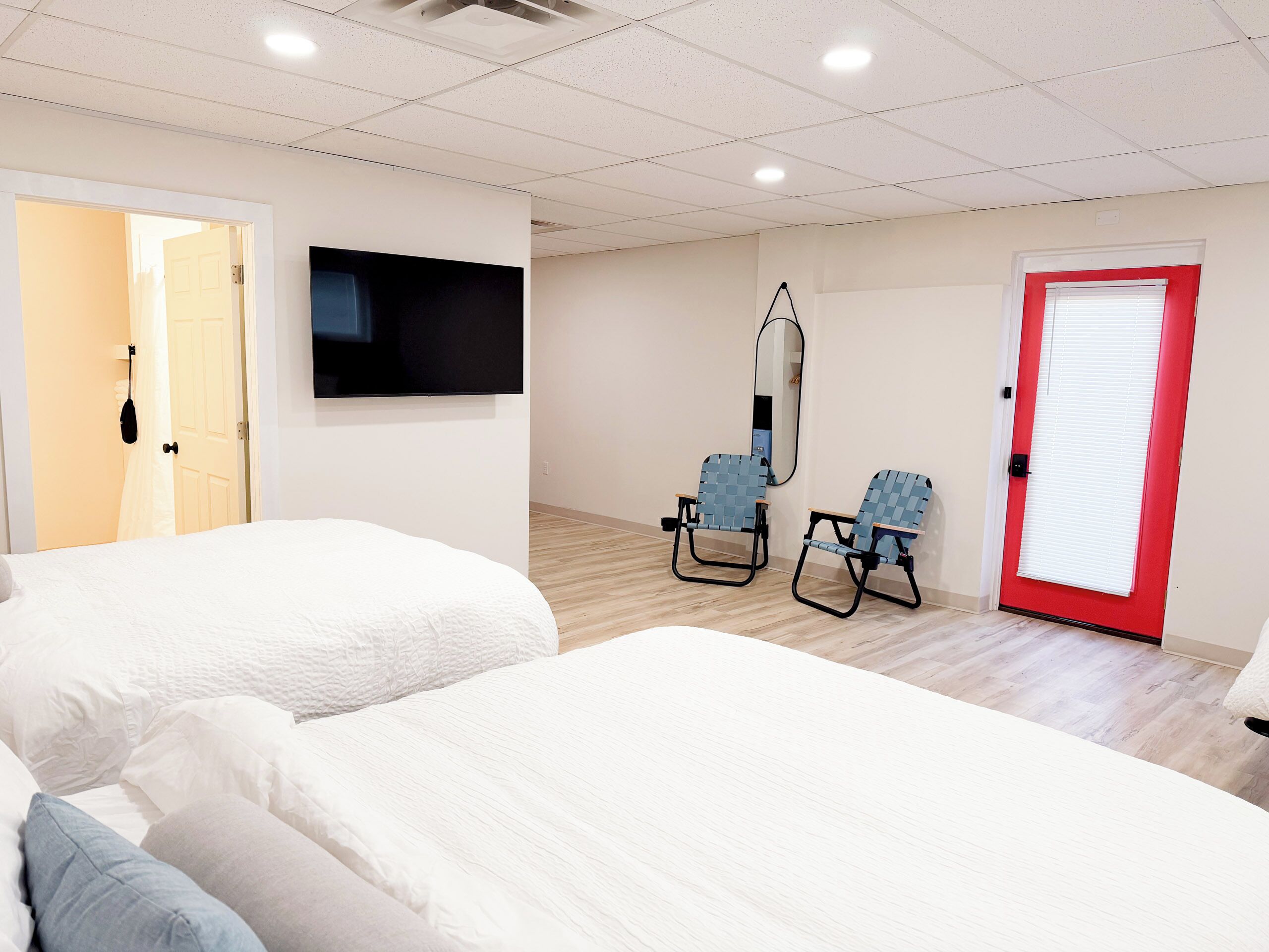 Quarto de hotel com duas camas, uma televisão, um espelho, duas cadeiras e uma porta vermelha.