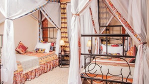 A room with two beds, each with a canopy. There are patterned bedspreads and pillows. A small table with a lamp is visible. - Jet Villas Entebbe (Entebbe)