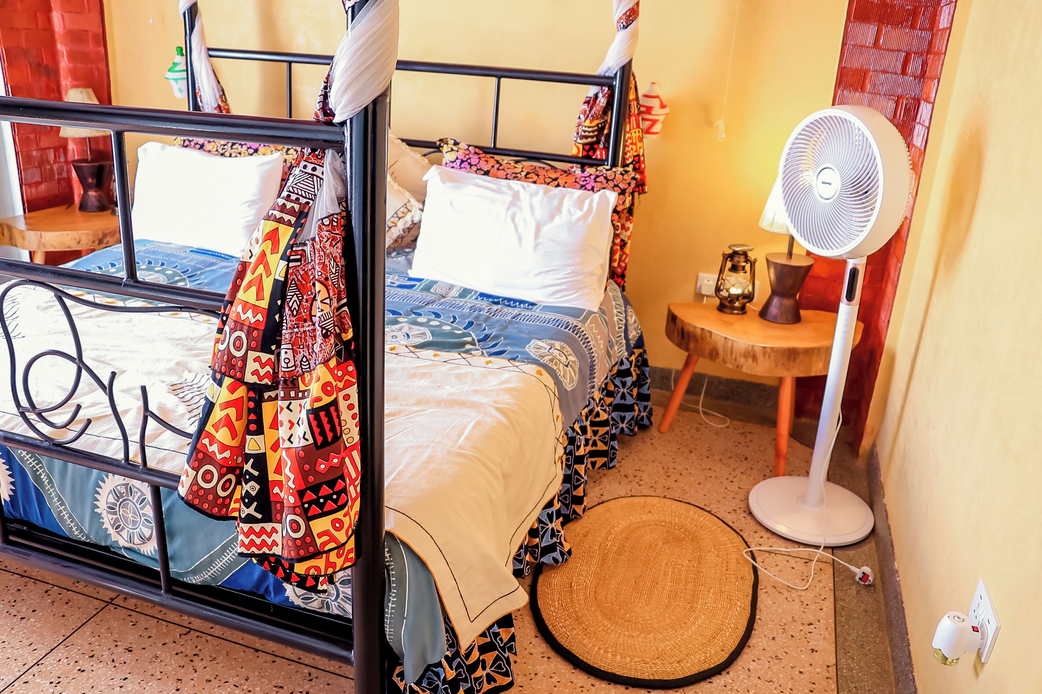A room with a bed, a standing fan, a small table, and a woven mat.
