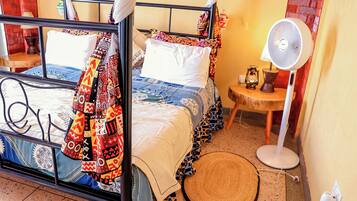 A room with a bed, a standing fan, a small table, and a woven mat.