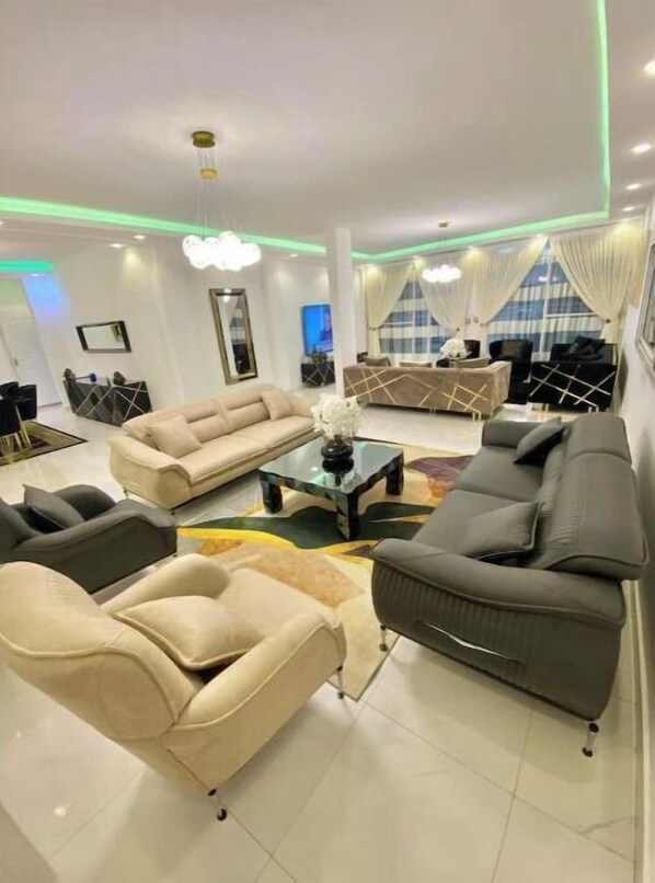 Un salon moderne avec un canapé beige, un canapé gris foncé et une table basse. On aperçoit un téléviseur au fond et une fenêtre avec des rideaux.