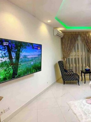 Un salon avec un grand téléviseur à écran plat fixé au mur, un fauteuil à motifs, une petite table d’appoint et un tapis blanc et moelleux.