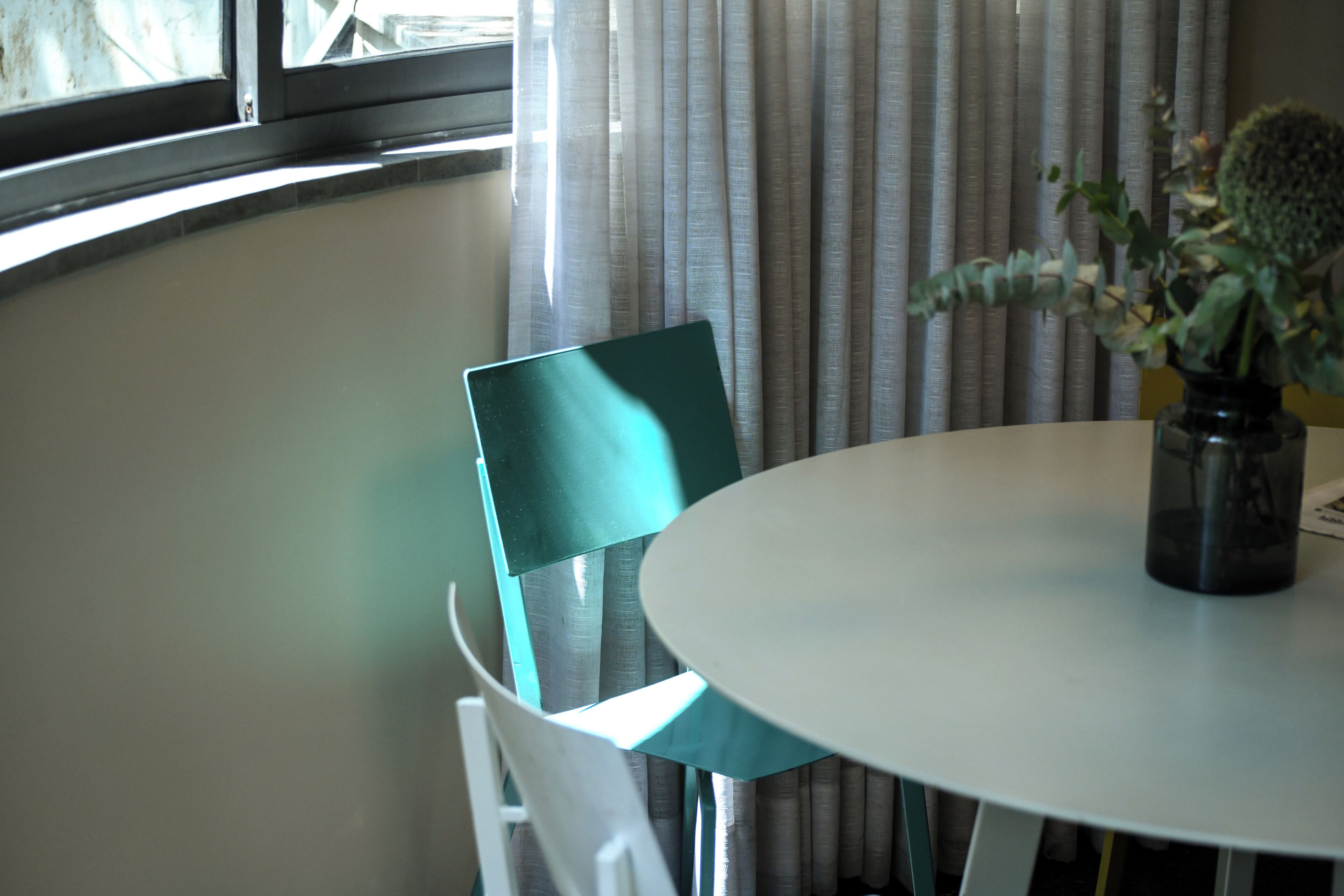 Ein Zimmer mit rundem Tisch, einem Stuhl, einer Vase mit Pflanze und einem Fenster mit Vorhängen.