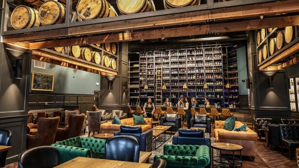 Un bar con barriles de madera, asientos de cuero y una estantería llena de botellas de licor.