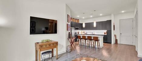 Una sala de estar moderna con una mesa de centro de madera, un televisor montado en la pared y una zona de cocina con taburetes de bar.