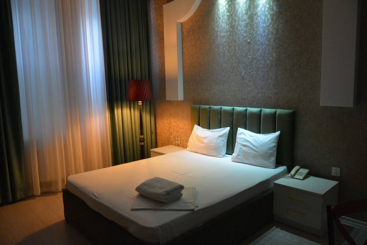 A hotel room with a bed, a bedside table with a phone, a nightstand, and a lamp.