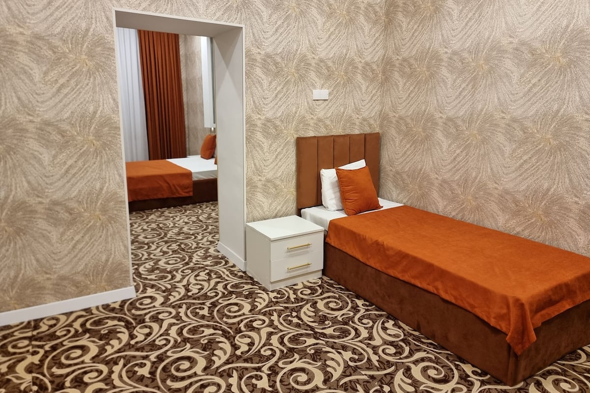 A hotel room with a patterned carpet, a bed with an orange bedspread, a bedside table with a white lamp, and a mirror reflecting the room.
