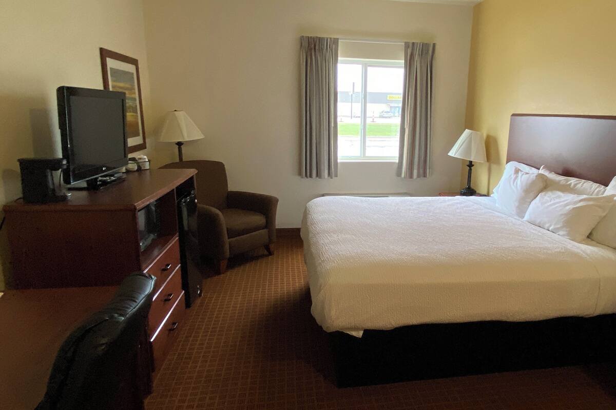 A hotel room with a bed, a desk with a television, a chair, a window with curtains, and a lamp.