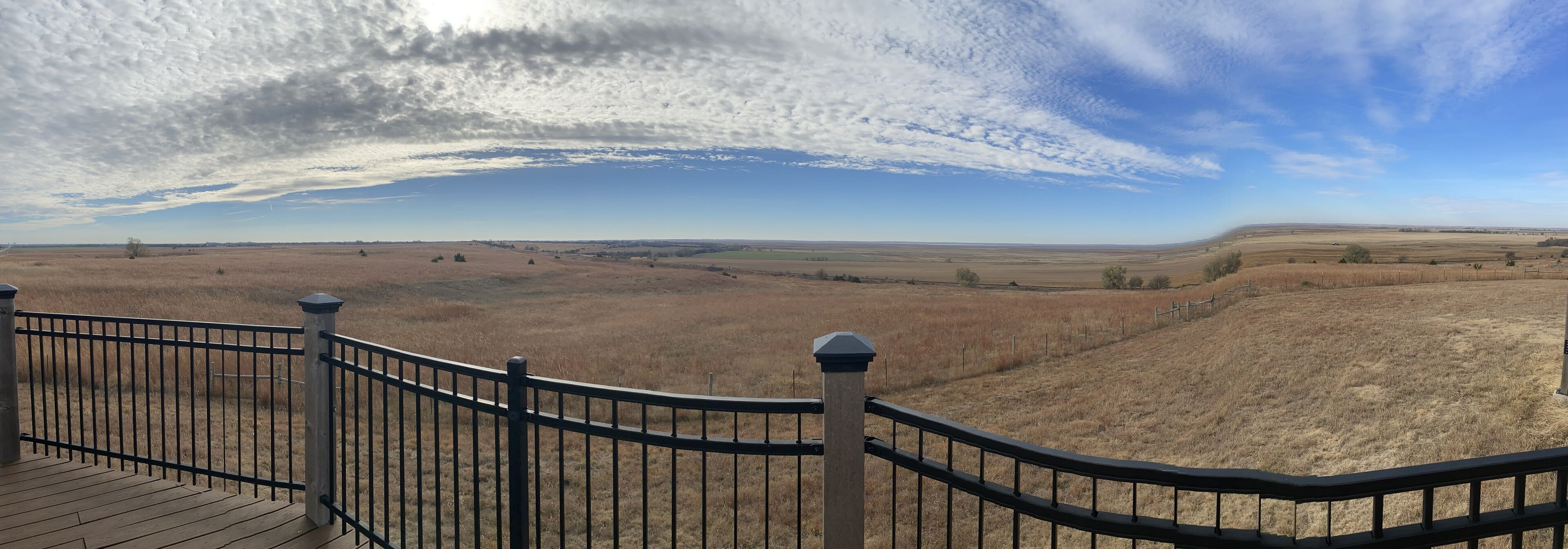 Une terrasse en bois, dotée d’une rambarde en métal, offre une vue imprenable sur un vaste paysage ouvert, avec des collines ondoyantes et un ciel dégagé.