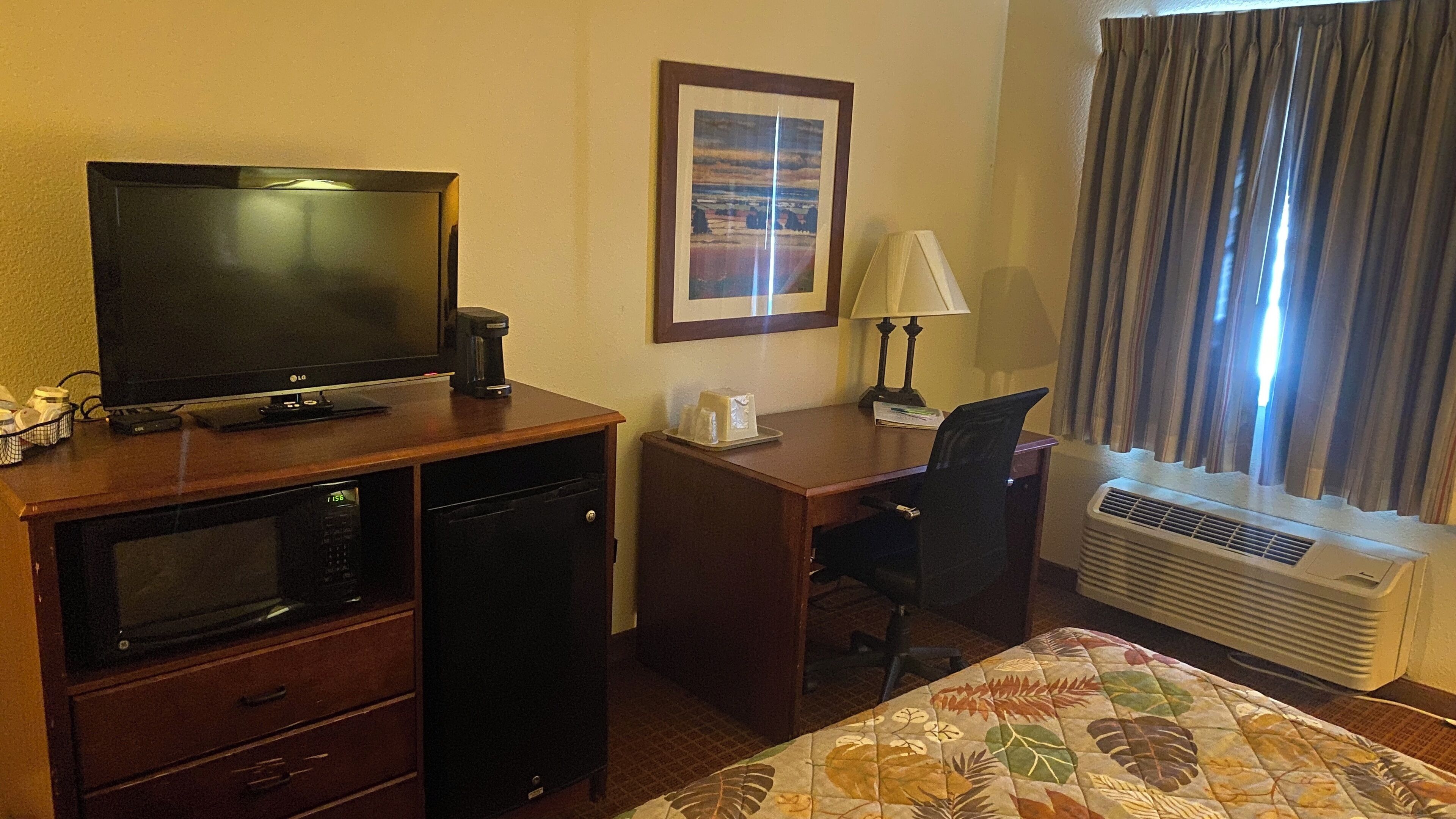 Hotelkamer met een flatscreen televisie, een koffiezetapparaat, een bureau met stoel, een inlijsting foto aan de muur en een raam met gordijnen.