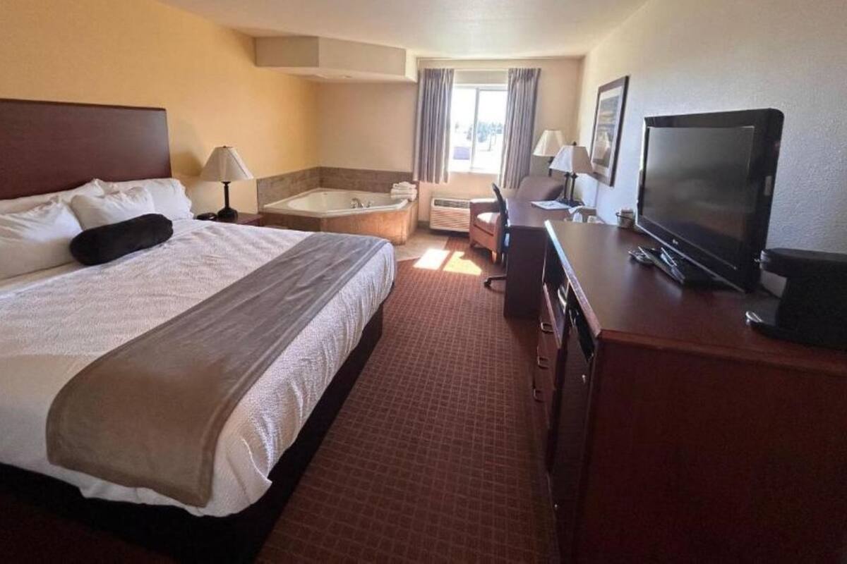 A hotel room with a large bed, a desk with a television, a chair, a bathtub, and a window with curtains.