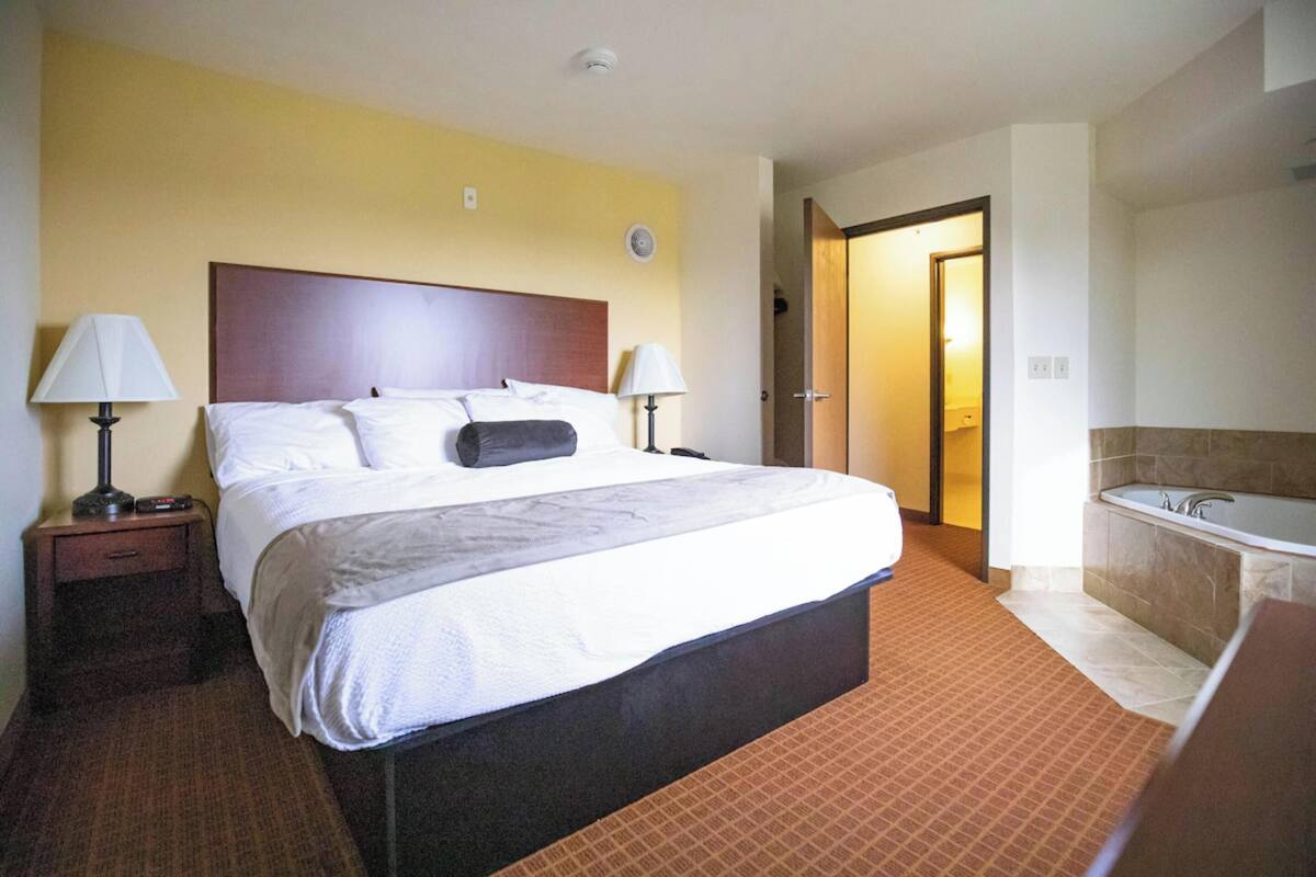 A hotel room with a large bed, two bedside tables with lamps, a bathroom with a bathtub, and a door leading to another room.