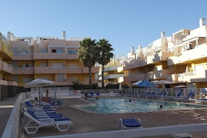 A pool area with lounge chairs and umbrellas, surrounded by multi-story buildings with balconies. - Marvelous T1 apartment - Cabanas de Tavira (Tavira)