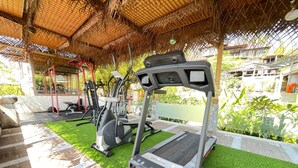 Un área de gimnasio al aire libre con equipo de ejercicio, incluyendo cintas de correr y máquinas de pesas, bajo un techo de paja.