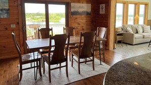 A wooden cabin interior with a dining table and chairs, a sofa, and a view of a lake through large windows. - Cabin on the Ocean-Carvers Cove (Vinalhaven)