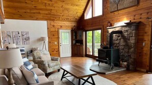 A cozy cabin interior with a stone fireplace, wooden floors, and a large window overlooking greenery. - Cabin on the Ocean-Carvers Cove (Vinalhaven)