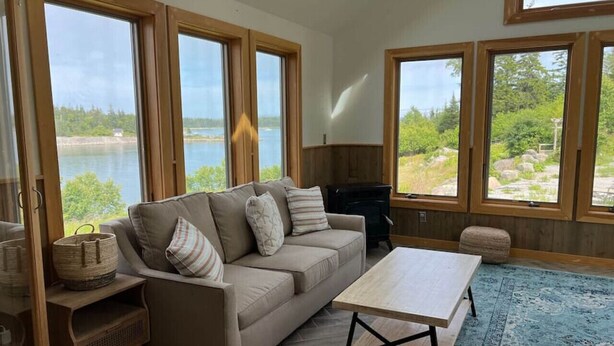 A living room with a sofa, a coffee table, and a view of a lake through large windows. - Cabin on the Ocean-Carvers Cove (Vinalhaven)