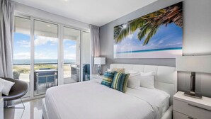A modern hotel room with a large bed, a balcony with a view of the beach, and a painting of a beach scene on the wall. - Sip of Sunshine Condo Oceanfront in Aruba (Noord)