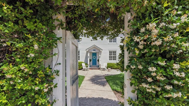 A pathway leading to a house with a blue door, surrounded by greenery and white walls. - Nostalgia of 1930’s with modern kitchen and baths, sleeps 8 (St Simons Island)