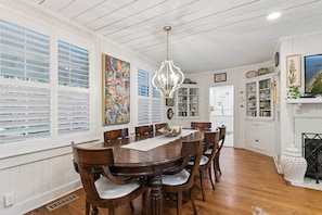 A dining room with a wooden table and chairs, a chandelier, and a large window with blinds.