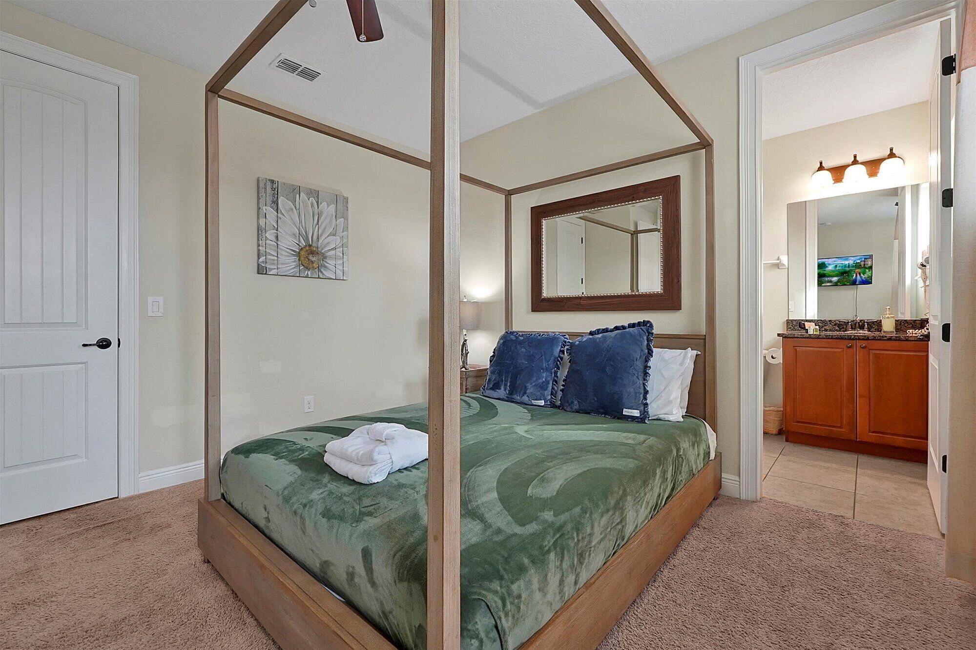 Un lit à baldaquin avec une couvre-lit verte, une serviette blanche et deux oreillers bleus, dans une pièce aux murs de couleur beige, au plafond en bois, et dont on aperçoit la salle de bain par une porte ouverte.