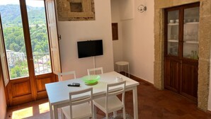 Zona pranzo con tavolo e sedie bianche, una televisione a muro e una parete in pietra con un armadio contenente bicchieri.
