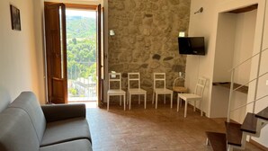 A living room with a grey sofa, a stone wall, a TV mounted on the wall, and a view of a balcony with chairs. - Three Bedroom Apartment (TROPEA)