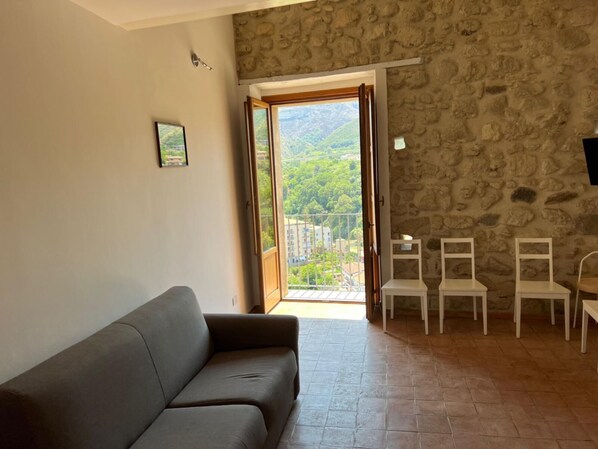 A living room with a grey sofa, a stone wall, and a balcony with a view of greenery and buildings.