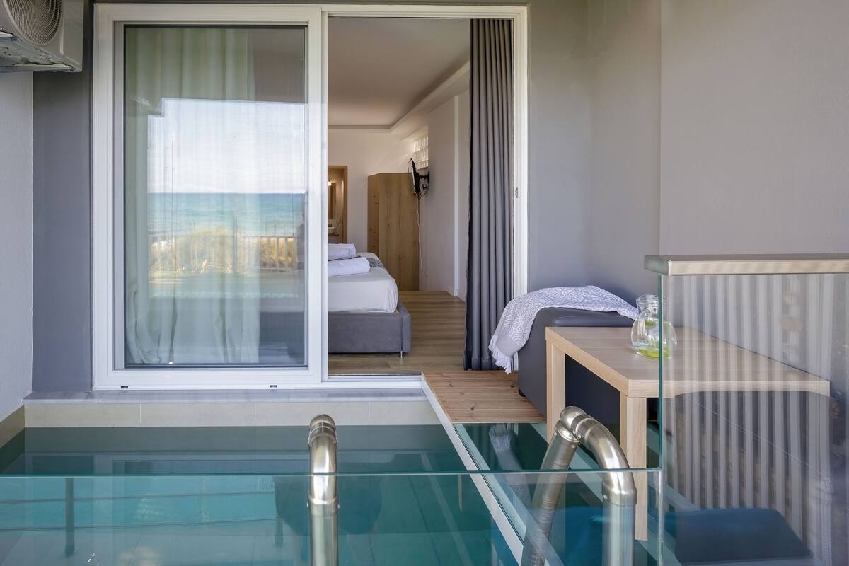Een moderne hotelkamer met een glazen zwembad, een houten vloer, een bed met witte lakens en een klein houten tafeltje met een glas water.